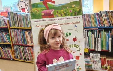 Dziewczynka w blond włosach trzyma swoją nową książkę, za nią stoi tablica z plakatami projektu Mała Książka Wielki Człowiek