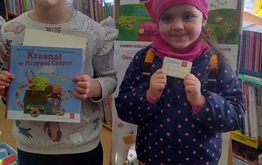 Stoją obok siebie dwie dziewczynki, jedna trzyma nową książkę, druga kartę Małego Czytelnika. Za nimi stoi tablica z plakatami projektu Mała Książka Wielki Człowiek