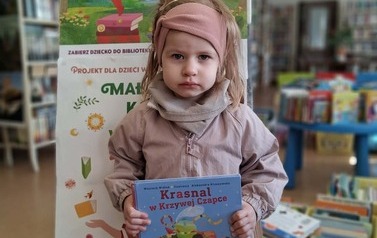 Dziewczynka w blond włosach trzyma swoją nową książkę, za nią stoi tablica z plakatami projektu Mała Książka Wielki Człowiek
