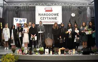 Grupa uczestnik&oacute;w Narodowego Czytania Dziad&oacute;w Adama Mickiewicza  stoi na scenie w strojach nawiązujących do dramatu. W tle napis "Narodowe Czytanie", a scenę ozdabiają dekoracje i rekwizyty nawiązujące do obrzędu dziad&oacute;w. Wydarzenie odbywa się w sali widowiskowej GOK