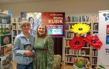 Czytelniczka z zakupioną książką stoi przy pisarce pozując. W tle duży ekran z wyświetlanym plakatem ze spotkania i dekoracje kwiatowe.