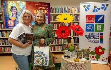 Pisarka i Czytelniczka pozują w bibliotece na tle regał&oacute;w z książkami.Jedna trzyma książki, druga papaierową torbę z prezentem. W tle znajduje się baner biblioteki oraz kolorowe dekoracje z kwiat&oacute;w.