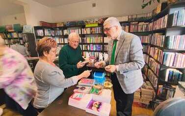 Mąż pisarki stoi przy biurku i sprzedaje seniorom książki pisarki.