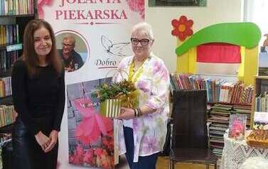 Bibliotekarka stoi obok pisarki trzymającej stroik jesienny. W tle baner promujący książkę Jolaanty Piekarskiej.