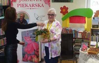 Bibliotekarka wręcza pisarce stroik jesienny w podziękowaniu za spotkanie. W tle baner promujący książkę pt " Warto. Naprawdę warto"