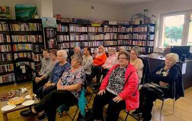 W bibliotece w Pinczynie siedzą na krzesłach seniorzy, z przodu stoi stolik z ciastem i kawą.