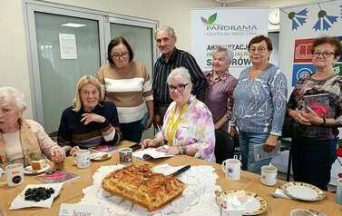 Uczestnicy spotkania siedzą i stoją wok&oacute;ł stołu z poczęstunkiem. Na stole znajduje się ciasto, fliżanki i talerzyki. W tle widoczny baner Centrum Szkolenia Panorama.