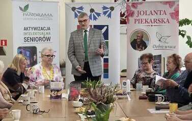 Przy stole pełnym czytelnik&oacute;w stoi mąż pisarki i przemawia. Na stole poczęstunek, a w tle  3 banery.