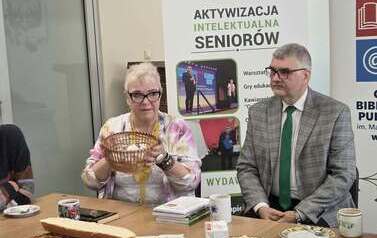 Pisarka siedzi obok męża przy stole z poczęstunkiem, trzyma w ręku koszyk, w kt&oacute;rym są na karteczkach ciekawe sentencje, myśli do losowania dla czytelnik&oacute;w. Za nimi baner promujący Centrum Szkolenia Panorama