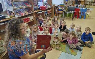 Dzieci siedzą na dywanie, a z przodu bibliotekarka czyta im bajkę mając książkę skierowaną obrazkami do dzieci
