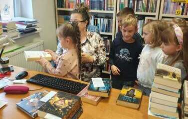Dzieci stoją przy biurku, bibliotekarka pomaga dziewczynce przyłożyć książkę do czytnika.