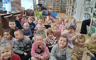 Cała grupa dzieci siedzi na dywanie w bibliotece.