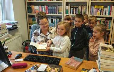 Dzieci stoją przy biurku, bibliotekarka pomaga dziewczynce przyłożyć książkę do czytnika.