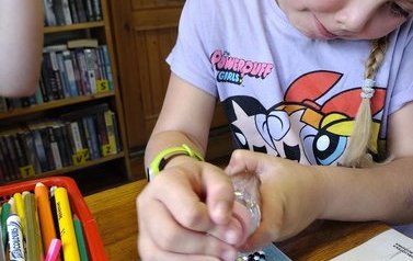Dziewczynka przy stole naciska buteleczkę z płynem i tworzy na plastikowej podstawce serduszka. Obok leżą kredki.