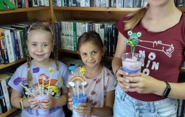 Trzy dziewczynki pokazują zrobione przez siebie kubeczki z kolorowym proszkiem, w kt&oacute;rych umieszczone są patyczki z wizerunkami tęczy, żabki, słoneczka i innych stwork&oacute;w.