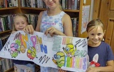 Trzy dziewczynki stoją i pokazują pokolorowany przez siebie bajkowy plakat na kt&oacute;rym widać Minionki.
