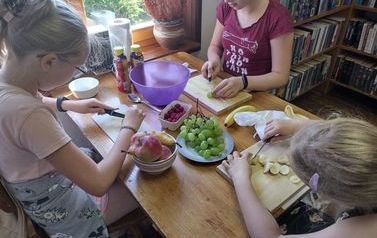 Trzy dziewczynki siedzą przy stoliku i kroją owoce do sałatki. Na stoliku leżą owoce, jogurty i miska.