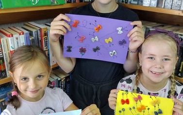 Trzy dziewczynki pokazują swoje prace na kartkach na kt&oacute;rych jest przyklejony kolorowy makaron w kształcie kokardek gdzie dorysowane są części tworząc obraz motylk&oacute;w
