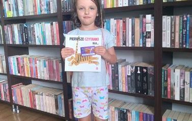 Dziewczynka stoi na tle p&oacute;łek z książkami trzymając w ręku książkę i kartę Małego Czytelnika