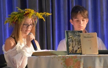 Dziewczyna i chlopak siedzą na scenie przy stole i dziewczyna czyta przez mikrofon. Na stole są postawione książki A. Mickeiwicza.