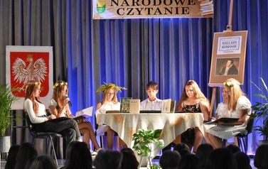 Scena z młodzieżą siedzącą przy stole i czytającą "Ballady i romanse" A. Mickiewicza