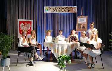 Młodzież biorąca udzial w czytaniu siedzi przy stoliku na scenie ubrana w na bialo i w wiankach na głowie. Stoi przy nich Kierownik biblioteki.