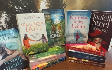 Nowe książki ułożone na stoliku z widocznymi tytułami. W tle pejzaż z zachodem słońca.