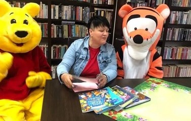 Pani siedzi przy stoliku w bibliotece z książeczką w ręku. Jest zwr&oacute;cona twarzą do Tygryska.