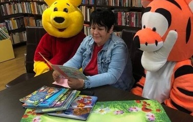 Pani czyta bajkę przy stoliku w bibliotece, obok siedzą Kubuś Puchatek i Tygrysek