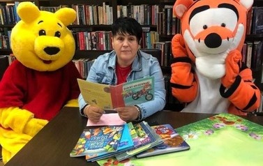 Pani siedzi przy stoliku w bibliotece trzymając w ręku bajkę, obok siedzą Kubuś Puchatek i Tygrysek