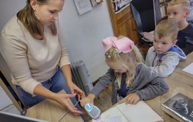 Troje dzieci stoi przy biurku i Pani bibliotekarka pokazuje pokolei jak działa czytnik do książek 