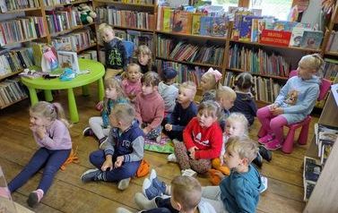 Dzieci siedzą na poduszkach na podłodze skierowane twarzą do bibliotekarki, kt&oacute;ra im czyta bajkę