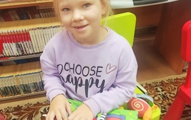 Dziewczynka o blond włosach siedzi na dywanie i przegląda poduszkową książeczkę, uśmiecha się.
