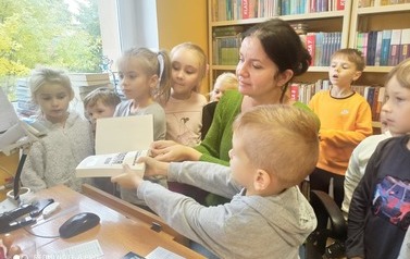 Dzieci stoją przy biurku obok Pani bibliotekarki. Jedno z dzieci- chłopiec przy pomocy bibliotekarki podsuwa książkę pod czytnik aby zeskanować kod kreskowy.