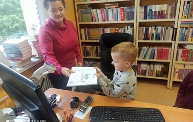 Chłopiec stoi przy biurku obok Pani bibliotekarki z książką i kieruje ją pod czytnik aby zapisała się w komputerze.