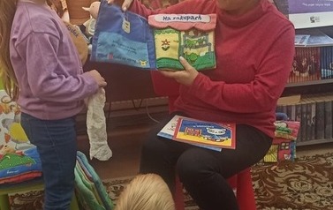 Dziewczynka stoi przy Pani bibliotekarce, w ręku trzyma siateczkę i bawi się w robienie zakup&oacute;w z książeczką pt. "Na zakupach" 