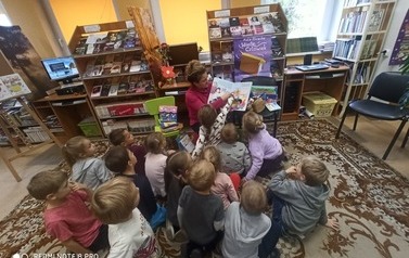 Dzieci siedzą na dywanie w bibliotece, po środku siedzi Pani bibliotekarka i czyta im bajkę.Pani i dziewczynka pokazują coś palcem w książce.