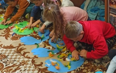 Dzieci siedzą na dywanie i przyklejają pi&oacute;rka do wyciętych z kartki skrzydeł 