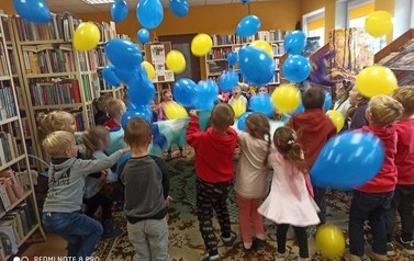 Dzieci stoją w k&oacute;łku, a nad nimi fruwają podrzucone kocem balony