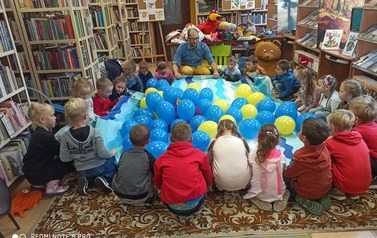 Prowadzący i dzieci siedzą na dywanie w k&oacute;łku, a po środku jest rozciągnięty koc zwypełniony balonami