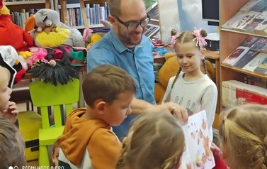 Troje dzieci stoi przed prowadzącym, kt&oacute;ry pokazuje ilustracje z książki dzieciom