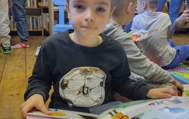 Chłopiec w ciemnej bluzce ogląda książeczkę. W tel widać inne dzieci.