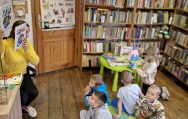 Dzieci siedzą na kocyku na podłodze, a Pani bibliotekarka siedzi na przecwko i  pokazuje im dwie książeczki