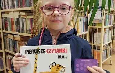 Dziewczynka pokazuje swoją nową książeczkę i kartę małego czytelnika. W tle jest kwiat doniczkowy i p&oacute;łki z książkami w bibliotece w Zblewie.