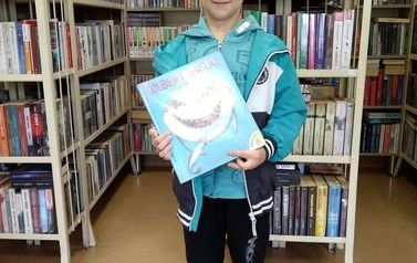 Chłopiec w niebieskiej kurtce pokazuje swoją wypożyczoną książkę. W tle p&oacute;łki z książkami w bibliotece w Zblewie