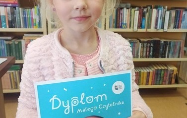 Dziewczynka o blond włosach pokazuje sw&oacute;j Dyplom Małego Czytelnika