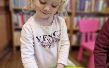 Dziewczynka o blond włosach stoi przy stoliczku i wykleja jeżyka z papieru.