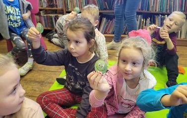 Dzieci siedzą w bibliotece na kocyku trzymając wyżej w rękach wycięte buźki.