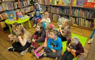 Dzieci siedzą w bibliotece na kocyku i słuchają bajki.