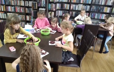 Dzieci siedzą w bibliotece przy stolikach i układają wzory na kartkach z kasztan&oacute;w.
